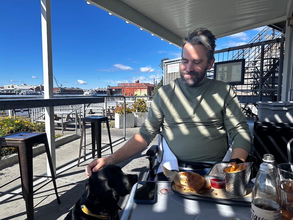 A man sits at a table with a meal, petting a black dog. The sky is blue, dock cranes and buildings are in the background.